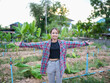 © buraratn - Young woman stands in lush garden, arms outstretched, embracing beauty of nature. She wears plaid shirt over black top and light gray pants, surrounded by green plants and serene landscape