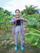 © buraratn - Young woman stands in lush green field, surrounded by banana plants, wearing plaid shirt and gray pants. She forms heart shape with her hands, expressing joy and positivity