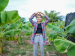 © buraratn - Young woman stands banana plantation, forming heart shape with her hands. She wears plaid shirt and gray pants, surrounded by lush green banana leaves. scene conveys joy and connection to nature