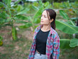 © buraratn - Young woman stands confidently in lush banana plantation, surrounded by vibrant green leaves. Her casual outfit, featuring plaid shirt over black top, complements serene rural setting