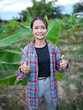 © buraratn - Young woman stands in banana plantation, smiling and giving thumbs up. She wears plaid shirt over black top and light gray pants, surrounded by lush green banana leaves