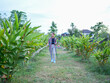 © buraratn - Woman stands in lush garden, watering plants with hose. She wears plaid shirt and light blue boots, surrounded by vibrant greenery and serene landscape