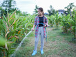 © buraratn - Young woman waters plants in lush green field using garden hose. She wears plaid shirt, black top, and gray pants, with blue rubber boots. serene environment reflects connection to nature