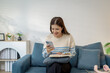 © PHOTO4U - A young woman sitting comfortably on a sofa holding a smartphone while smiling and reading a message, with an open notebook resting on her lap in a bright cozy room.