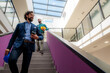© DusanJelicic - Businessman holding takeaway coffee and safety helmet walking downstairs in modern office building, followed by an engineer holding a yellow safety helmet