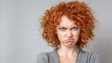 A woman with curly red hair, wearing a gray sweater, making a face with her mouth open and eyes narrowed, against a plain background.