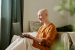 © AnnaStills - Caucasian young adult woman with alopecia sitting on bed using laptop smiling, natural light illuminating face, relaxed posture, short hairless scalp clearly visible