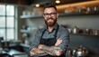 © miss irine - Smiling bearded male chef with glasses, arm tattoos, stands confidently in modern kitchen. Wears grey shirt, black apron, crossing arms. Happy professional cook looks friendly, ready for culinary