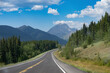 © Connect Images - Winding road through lush green forest with mountains in the background on a sunny day. Waterton Lakes National Park, Alberta, Canada