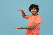 © Prostock-studio - A cheerful young Indian man with curly hair in a pink t-shirt smiles at the camera while holding something invisible between his hands. He stands against a bright blue studio backdrop.