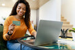 © Prostock-studio - A woman sits at her desk in a bright workspace, happily making a purchase online. She holds a credit card in one hand while focusing on her laptop screen.