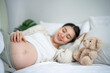 © Kawee - Asian young pregnant woman gently stroking belly while lying down on bed.