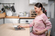 © Kawee - Asian young pregnant businesswoman using laptop computer in kitchen.