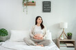 © Kawee - Asian young pregnant woman gently stroking belly while sitting on bed.