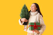 © Pixel-Shot - Young woman with small Christmas tree and gift box on yellow background