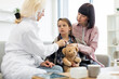 © sofiko14 - A female doctor uses a stethoscope to listen to a young girl's heart while her mother comforts her. The girl holds a teddy bear.
