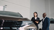 © wedmoments.stock - A car dealer and a customer discuss the features of a new car at a showroom, demonstrating the buying process and customer service experience in a comfortable environment.
