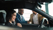 © wedmoments.stock - A smiling woman in a car at a car dealership watches a handshake between men, showing the excitement of buying a new car in a modern automotive environment.