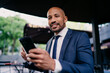 © BullRun - Cheerful businessman in formal suit smiling at camera, holding smartphone and coffee, portraying approachability, success and digital comfort in modern mobile workspace.