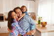© Stockphotodirectors - A happy family is capturing a joyful moment as they take a selfie in their bright kitchen. The parents are smiling, and their child is playfully perched on one parent's back.
