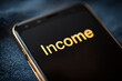 © kalafoto - Close-up of 'Income' text displayed in golden letters on a dark smartphone screen, symbolizing mobile finance.