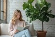 © stockphoto02 - Young woman reads book on sofa. Large fiddle leaf fig plant beside her. Cozy, well-lit room with white brick wall.