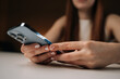 © dikushin - Close-up cropped shot of unrecognizable woman holding modern smartphone, browsing internet or using social media apps, enjoying mobile technology sitting at white table in dark comfortable setting.