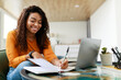 © Prostock-studio - A young woman is happily engaged in her studies at home. She is taking notes while seated at a glass table, with her laptop in front of her and natural light illuminating the room.