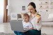 © Prostock-studio - A woman is sitting on the floor with her young child beside her. She holds a yellow credit card while browsing on a laptop in a comfortable living space.