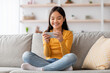 © Prostock-studio - A girl sits cross-legged on a couch, smiling as she plays games on her smartphone. The cozy living room features light decor and a relaxed atmosphere.