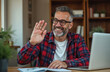 © Vadym - Mature man waving hello during remote work video call on laptop. He smiles, wearing glasses and a plaid shirt. Man works from home office, using computer, engaging in online communication.
