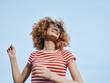 © SHOTPRIME STUDIO - Young person with curly hair smiles under a clear blue sky, wearing a red and white striped shirt and sunglasses, radiating carefree summer energy and playful confidence.