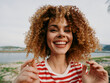© SHOTPRIME STUDIO - Joyful portrait of a young woman with curly hair smiling broadly outdoors by a lake, wearing a red and white striped shirt, exuding vibrant energy.