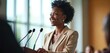 © Viktor - Smiling Black woman delivers speech at podium during business conference. Stands confidently with microphones. Female speaker engages large audience, shares pro insights at event. Leader presents
