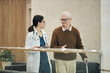 © DragonImages - Middle aged Asian woman doctor walking and talking with senior Caucasian man holding cane in medical facility, both engaged in conversation, doctor holding digital tablet