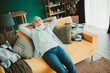 © deagreez - Relaxed senior man lounges on a bright orange sofa enjoying rest and casual comfort at home in a cozy living room with cushions