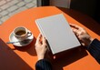 © 2Blurry - Woman's hands holding a blank white book cover at a sunlit cafe table with espresso