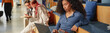 © Seventyfour - Young adult woman sitting on bleachers using laptop, focusing on screen while two young adult people in background talking and reading book, modern educational setting
