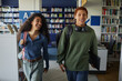 © Seventyfour - Teenage Biracial girl and boy walking together in library, both carrying backpacks and smiling, boy holding laptop and headphones around neck, bookshelves in background