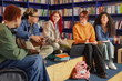 © Seventyfour - Group of diverse teenagers sitting together in library discussing schoolwork, multiethnic boys and girls interacting with notebooks and laptops, bookshelves in background