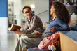 © Seventyfour - Asian young adult man smiling and holding book while talking to biracial young adult woman sitting beside him, both engaging in conversation with laptop and backpack visible