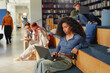 © Seventyfour - Young adult woman sitting on steps using laptop, surrounded by diverse group of young adults and teenagers studying with digital devices in modern library setting
