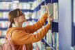 © Seventyfour - Caucasian young adult woman with glasses reaching for book on library shelf, wearing backpack, standing in front of rows of books, engaging in academic or leisure reading activity