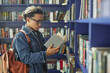 © Seventyfour - Young adult Asian man wearing glasses standing in library reading book while carrying backpack, focusing on pages with shelves of books in background, engaging in studying or research
