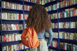 © Seventyfour - Black teenage girl standing with backpack in front of bookshelves in library, facing away from camera, long curly hair visible, exploring book selection, casual clothing