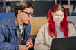 © Seventyfour - Young adult Asian man and young adult Caucasian woman sitting together using laptop, man holding wooden blocks, both focusing on screen, collaborating on project or studying