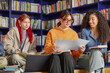 © Seventyfour - Three young adult women, including Caucasian and biracial individuals, sitting together in library holding papers and notebooks, collaborating on academic project, smiling and studying