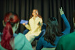 © Seventyfour - Teenage girl with brown hair sitting on stage holding tablet and remote, speaking to audience while teenage girl with dark hair raising hand in foreground, classroom or seminar setting