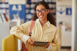 © Seventyfour - Portrait of young adult biracial woman smiling while holding digital tablet and wearing eyeglasses in library setting, standing with backpack and looking directly at camera