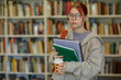 © Seventyfour - Portrait of Caucasian young adult woman with red hair and glasses standing in library holding notebooks and coffee cup, wearing casual clothing, looking directly at camera